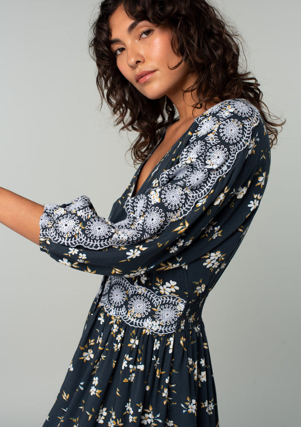 [Color: Navy/Powder Blue] A close up side facing image of a brunette model wearing a bohemian navy blue mini dress with a yellow and white floral print. With lace trimmed long sleeves, a button front, and a lace trimmed waist. 