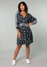 [Color: Navy/Powder Blue] A full body front facing image of a brunette model wearing a bohemian navy blue mini dress with a yellow and white floral print. With lace trimmed long sleeves, a button front, and a lace trimmed waist. 