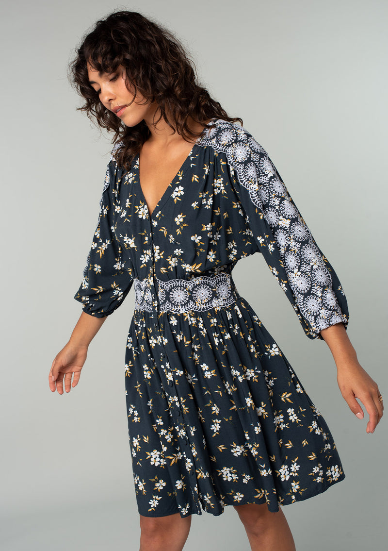 [Color: Navy/Powder Blue] A front facing image of a brunette model wearing a bohemian navy blue mini dress with a yellow and white floral print. With lace trimmed long sleeves, a button front, and a lace trimmed waist. 