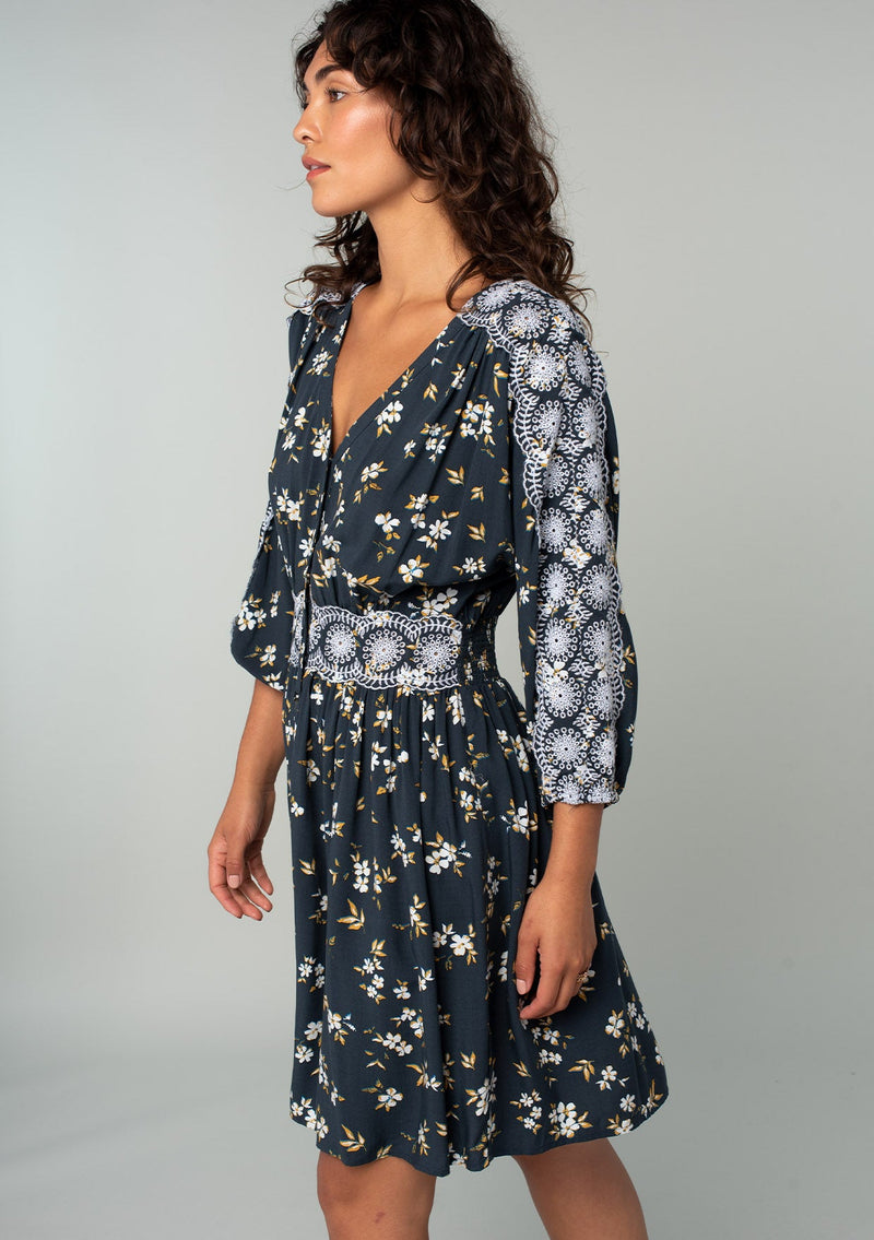 [Color: Navy/Powder Blue] A side facing image of a brunette model wearing a bohemian navy blue mini dress with a yellow and white floral print. With lace trimmed long sleeves, a button front, and a lace trimmed waist. 