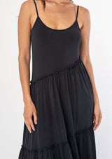 [Color: Black] A woman wearing a soft and stretchy black knit tank mid length dress with spaghetti straps and ruffled trim. 