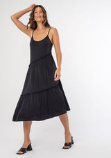 [Color: Black] A woman wearing a soft and stretchy black knit tank mid length dress with spaghetti straps and ruffled trim. 