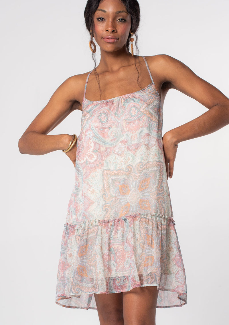 [Color: Blush/Aqua] A model wearing a flowy bohemian mini tank dress in a pink and blue paisley print. A sheer chiffon mini dress with spaghetti straps and a cross strap back detail. 