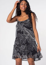[Color: Black/Ivory] A model wearing a bohemian black flowy chiffon mini dress with gold metallic details, a palm print, and adjustable spaghetti straps. 