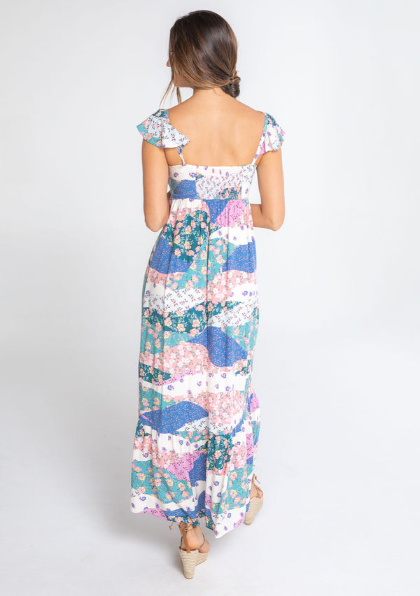 [Color: Ivory/Jade] A model wearing a white, pink, and blue floral patchwork print maxi dress with a sweetheart neckline, short flutter sleeves, and an empire waist. 