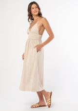 [Color: Natural] A model wearing a bohemian sleeveless mid length dress in a natural linen stripe, with tie shoulder strap detail. 