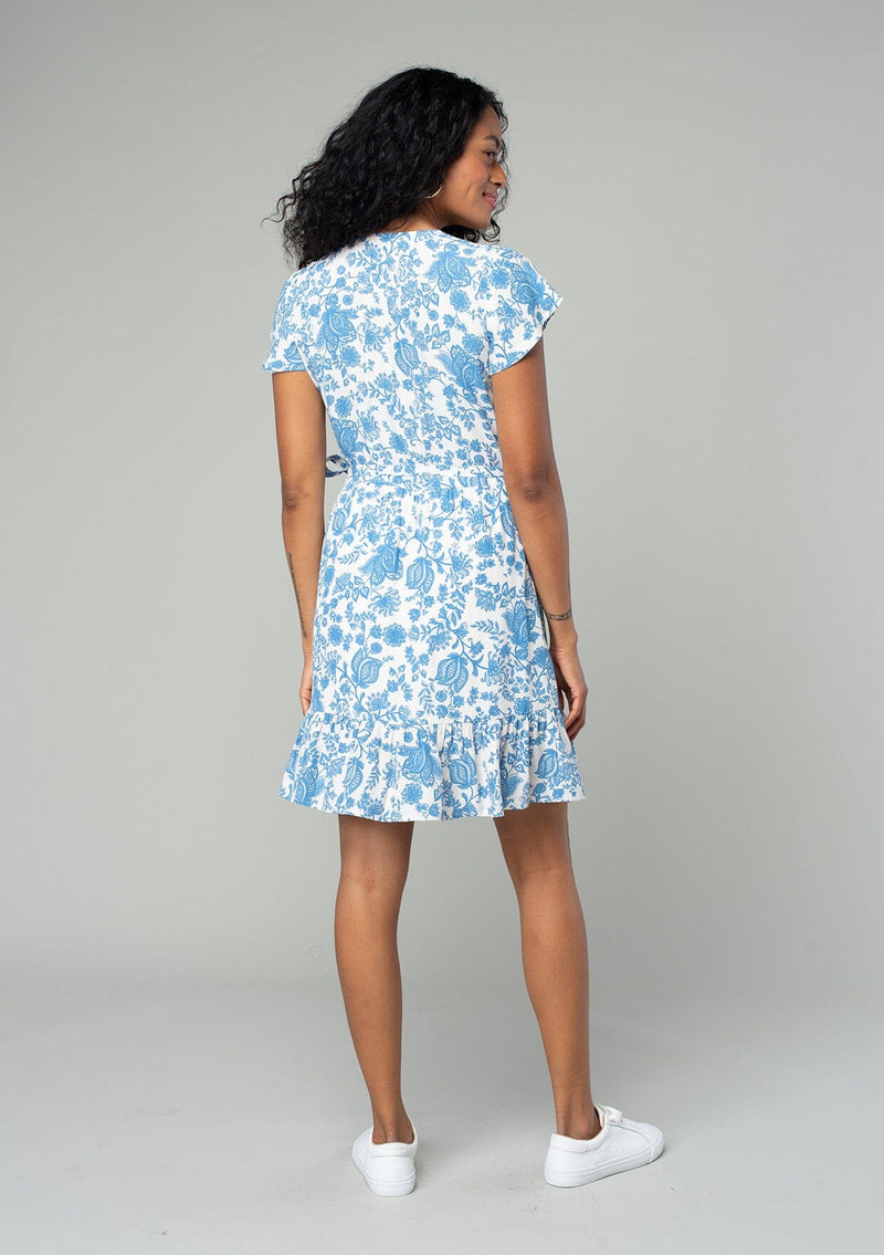 [Color: Cream/Dusty Blue] A back facing image of a brunette model wearing a classic mini wrap dress in a white and blue floral print. With short flutter sleeves, a side tie wrap closure, and a ruffled hemline. 