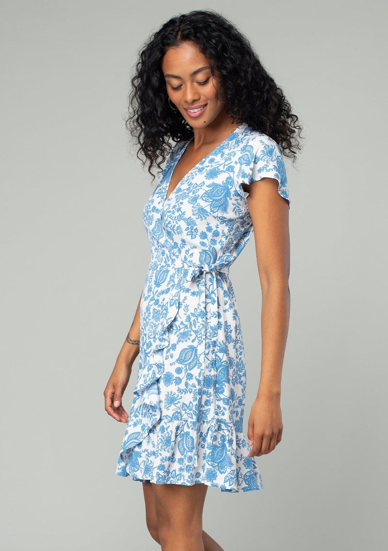 [Color: Cream/Dusty Blue] A side facing image of a brunette model wearing a classic mini wrap dress in a white and blue floral print. With short flutter sleeves, a side tie wrap closure, and a ruffled hemline. 
