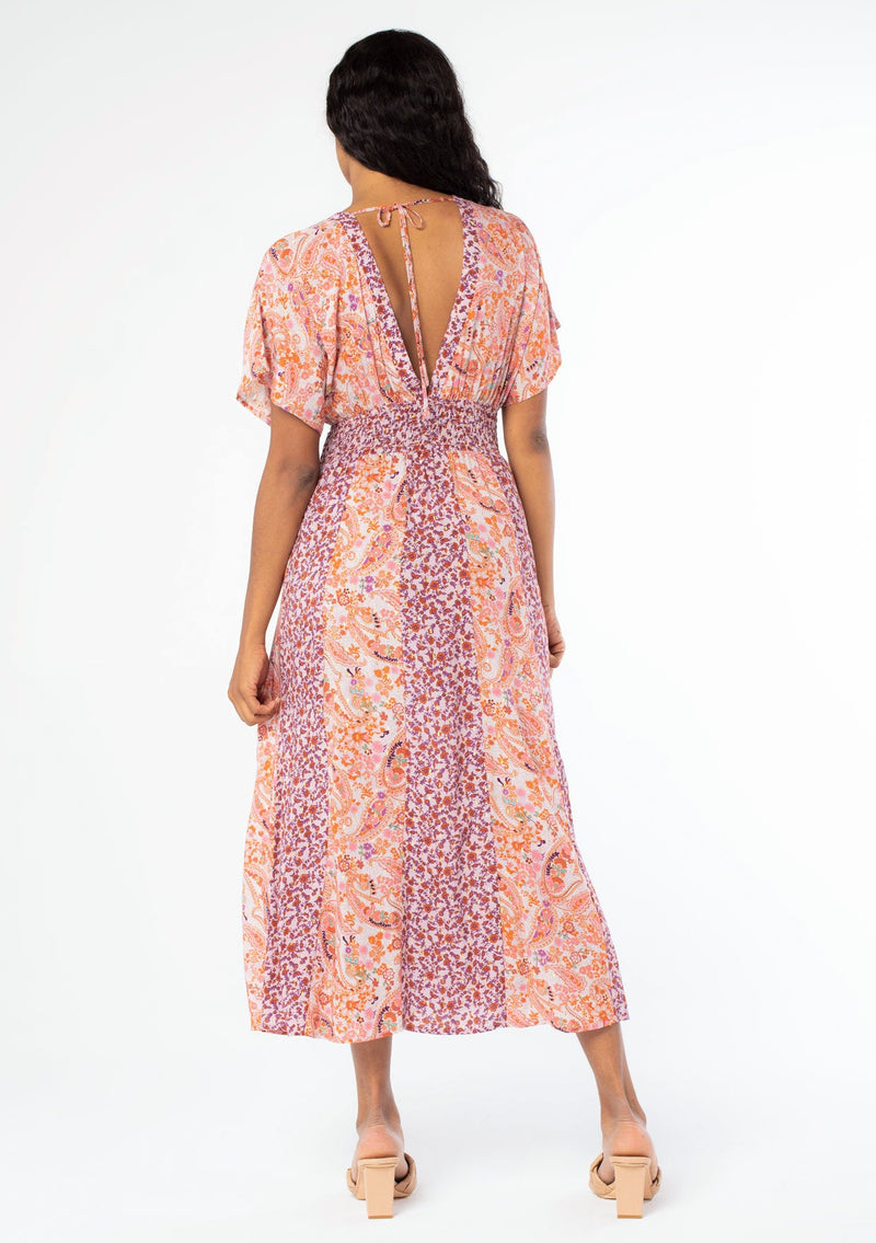 [Color: Lavender/Orange] A full back image of a black model with long dark wavy hair wearing a purple and orange mixed floral print maxi dress with short kimono sleeves, a smocked elastic waist, and a side slit. 