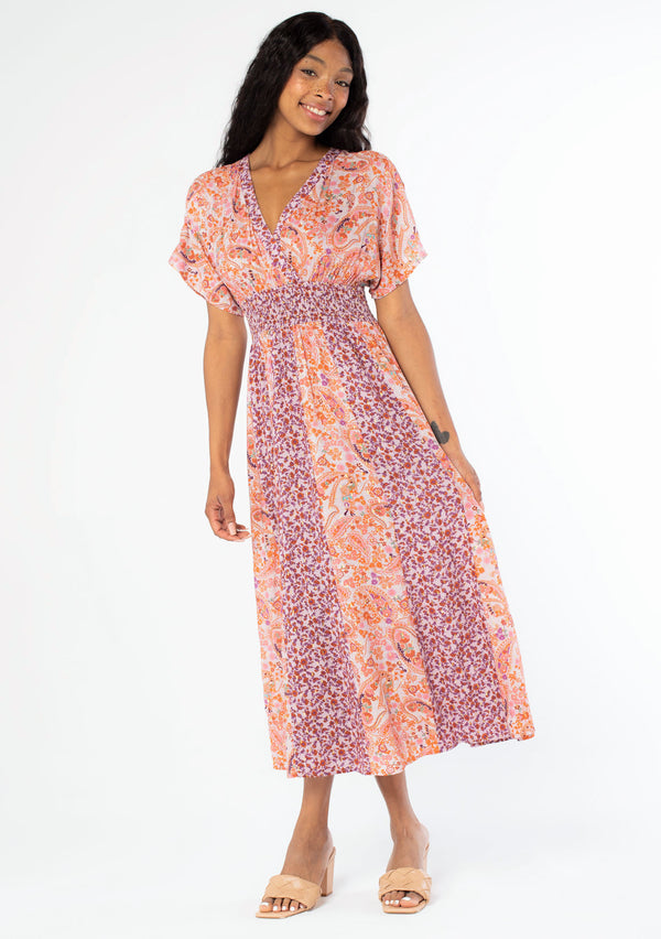 [Color: Lavender/Orange] A front facing image of a black model with long dark wavy hair wearing a purple and orange mixed floral print maxi dress with short kimono sleeves, a smocked elastic waist, and a side slit. 