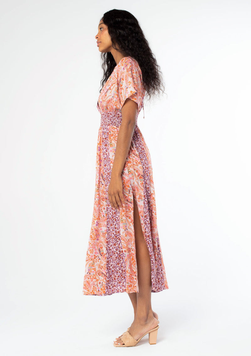 [Color: Lavender/Orange] A side image of a black model with long dark wavy hair wearing a purple and orange mixed floral print maxi dress with short kimono sleeves, a smocked elastic waist, and a side slit. 