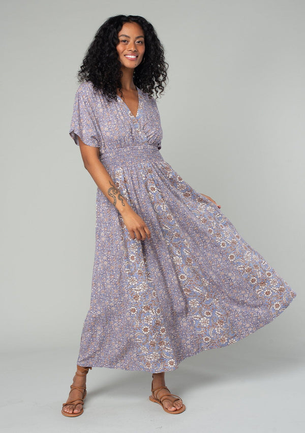 [Color: Grey/Natural] A front facing image of a brunette model wearing a flowy bohemian resort maxi dress in a grey and purple mixed floral print. With short kimono sleeves, an open back with tie closure, a smocked elastic waist, and a side slit. 