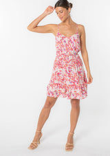 [Color: Blush/Red] A model wearing a pretty pink floral sleeveless mini dress with a ruffled tiered skirt and a spaghetti strap cross back detail. 