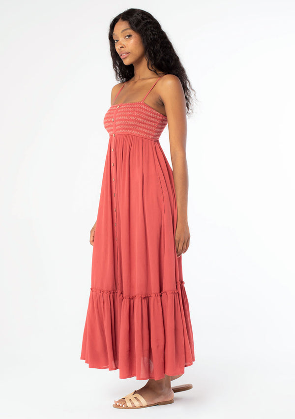[Color: Burnt Sienna] A woman wearing a rust red crinkle gauze sleeveless button front maxi dress with smocked bust and adjustable spaghetti straps.