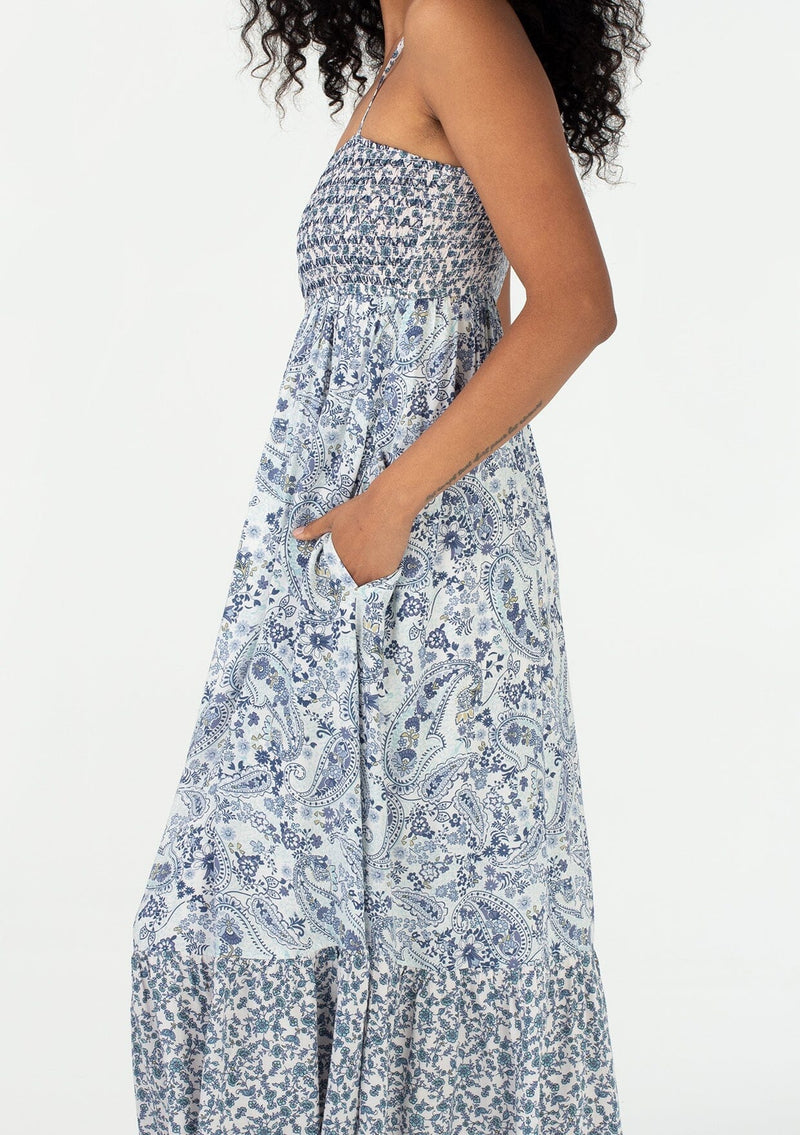 [Color: Ivory/Blue] A close up side facing image of a brunette model wearing a bohemian spring resort maxi dress in an ivory and blue mixed floral and paisley print. With adjustable spaghetti straps, a straight neckline, a slim fit smocked bodice, a button front, a flowy tiered skirt, and side pockets. 