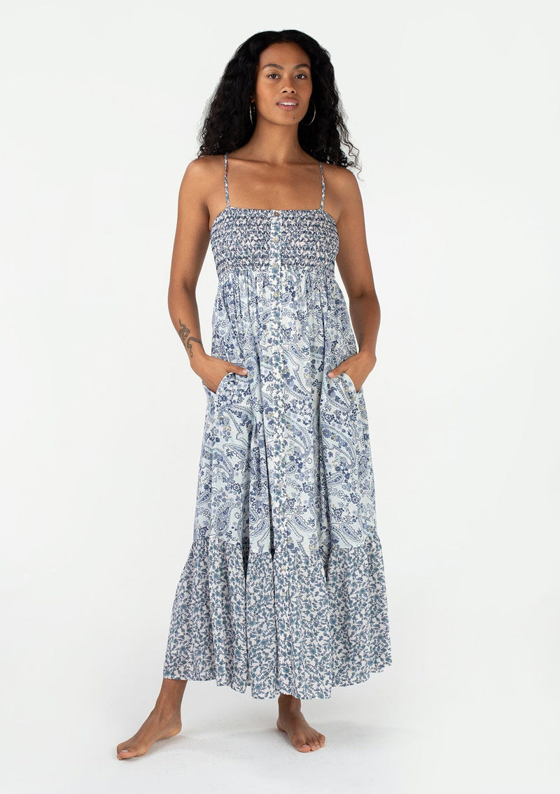 [Color: Ivory/Blue] A front facing image of a brunette model wearing a bohemian spring resort maxi dress in an ivory and blue mixed floral and paisley print. With adjustable spaghetti straps, a straight neckline, a slim fit smocked bodice, a button front, a flowy tiered skirt, and side pockets. 