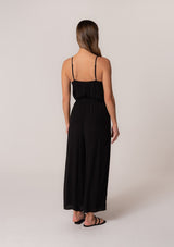 [Color: Black] A back facing image of a brunette model wearing a solid black bohemian one piece jumpsuit. With adjustable spaghetti straps, a scoop neckline, an elastic waist, an adjustable braided rope belt with tassel accents, and a long wide leg.