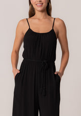[Color: Black] A close up front facing image of a brunette model wearing a solid black bohemian one piece jumpsuit. With adjustable spaghetti straps, a scoop neckline, an elastic waist, an adjustable braided rope belt with tassel accents, and a long wide leg.