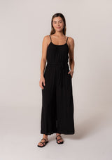 [Color: Black] A front facing image of a brunette model wearing a solid black bohemian one piece jumpsuit. With adjustable spaghetti straps, a scoop neckline, an elastic waist, an adjustable braided rope belt with tassel accents, and a long wide leg.