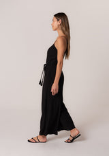 [Color: Black] A side facing image of a brunette model wearing a solid black bohemian one piece jumpsuit. With adjustable spaghetti straps, a scoop neckline, an elastic waist, an adjustable braided rope belt with tassel accents, and a long wide leg.