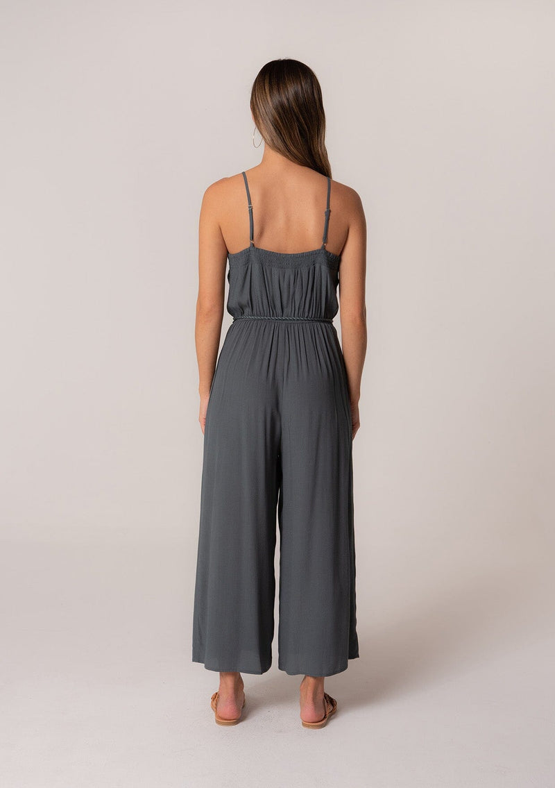 [Color: Slate] A back facing image of a brunette model wearing a solid blue bohemian one piece jumpsuit. With adjustable spaghetti straps, a scoop neckline, an elastic waist, an adjustable braided rope belt with tassel accents, and a long wide leg.