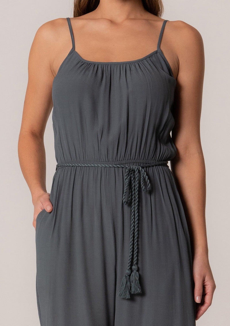 [Color: Slate] A close up front facing image of a brunette model wearing a solid blue bohemian one piece jumpsuit. With adjustable spaghetti straps, a scoop neckline, an elastic waist, an adjustable braided rope belt with tassel accents, and a long wide leg. 