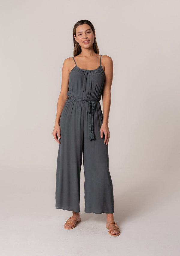[Color: Slate] A front facing image of a brunette model wearing a solid blue bohemian one piece jumpsuit. With adjustable spaghetti straps, a scoop neckline, an elastic waist, an adjustable braided rope belt with tassel accents, and a long wide leg.