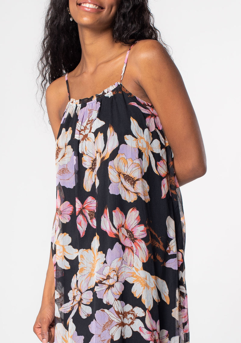 [Color: Black/Lilac] A model wearing a black bohemian maxi tank dress in a purple floral print. A sheer chiffon maxi dress with spaghetti straps and a flowy silhouette. 
