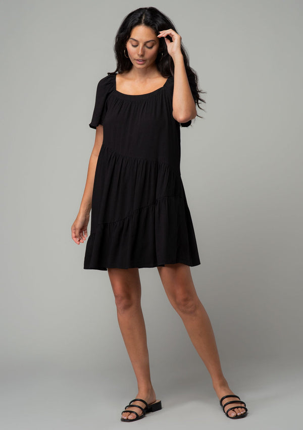 [Color: Black] A full body front facing image of a brunette model wearing a black linen blend mini dress. A flowy bohemian mini dress with short sleeves, a wide square neckline, and an asymmetric tiered hemline. With an open back detail. 