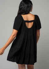 [Color: Black] A back facing image of a brunette model wearing a black linen blend mini dress. A flowy bohemian mini dress with short sleeves, a wide square neckline, and an asymmetric tiered hemline. With an open back detail. 