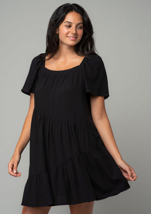 [Color: Black] A half body front facing image of a brunette model wearing a black linen blend mini dress. A flowy bohemian mini dress with short sleeves, a wide square neckline, and an asymmetric tiered hemline. With an open back detail. 