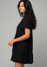 [Color: Black] A side facing image of a brunette model wearing a black linen blend mini dress. A flowy bohemian mini dress with short sleeves, a wide square neckline, and an asymmetric tiered hemline. With an open back detail. 