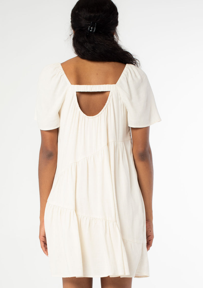 [Color: Natural] A back facing image of a black model wearing a natural, off white linen blend mini dress. A flowy bohemian mini dress with short sleeves, a wide square neckline, and an asymmetric tiered hemline. With an open back detail. 