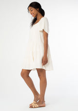 [Color: Natural] A full body side facing image of a black model wearing a natural, off white linen blend mini dress. A flowy bohemian mini dress with short sleeves, a wide square neckline, and an asymmetric tiered hemline. With an open back detail. 