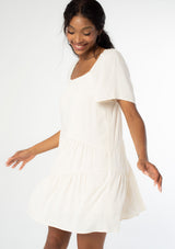 [Color: Natural] A side facing image of a black model wearing a natural, off white linen blend mini dress. A flowy bohemian mini dress with short sleeves, a wide square neckline, and an asymmetric tiered hemline. With an open back detail. 