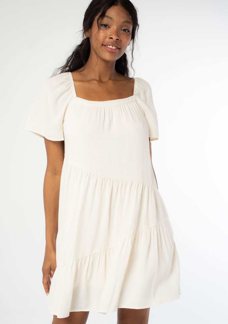 [Color: Natural] A front facing image of a black model wearing a natural, off white linen blend mini dress. A flowy bohemian mini dress with short sleeves, a wide square neckline, and an asymmetric tiered hemline. With an open back detail. 