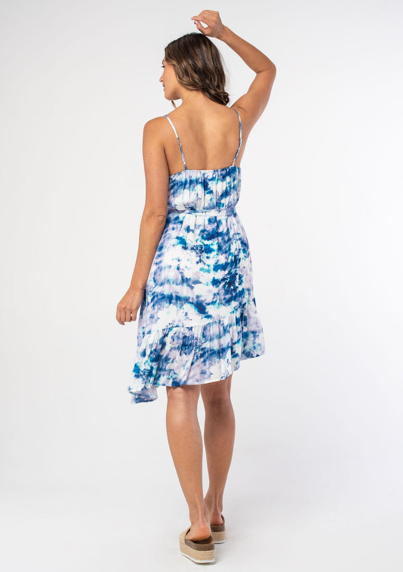 [Color: Navy/Teal] A woman wearing a blue and white watercolor floral print sleeveless mini dress with spaghetti straps and a ruffled asymmetric hemline. 