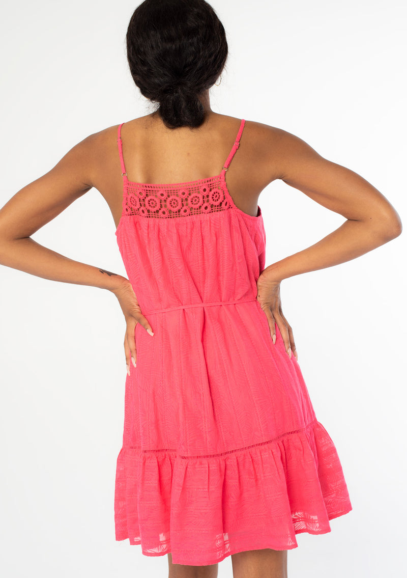 [Color: Watermelon] A back facing image of a black model wearing a bright pink bohemian sleeveless mini tank dress with spaghetti straps, a crochet knit trim, and a tassel tie waist belt. 