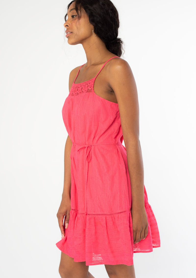 [Color: Watermelon] A side facing image of a black model wearing a bright pink bohemian sleeveless mini tank dress with spaghetti straps, a crochet knit trim, and a tassel tie waist belt. 