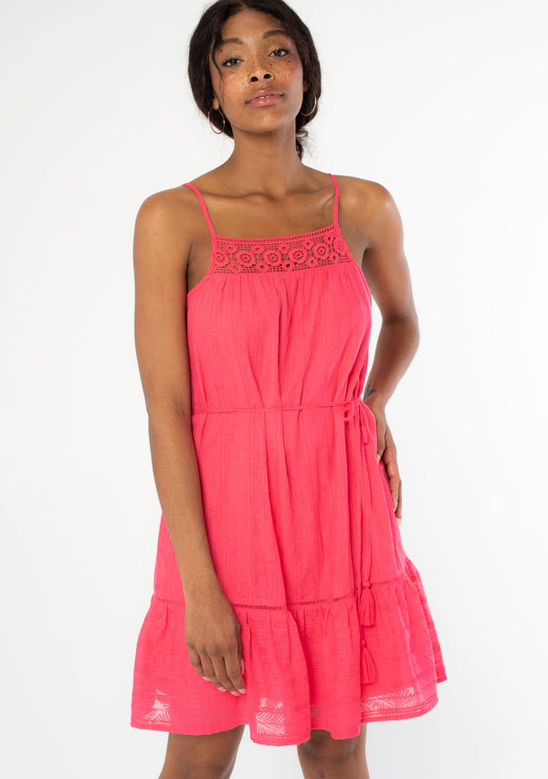 [Color: Watermelon] A front facing image of a black model wearing a bright pink bohemian sleeveless mini tank dress with spaghetti straps, a crochet knit trim, and a tassel tie waist belt. 