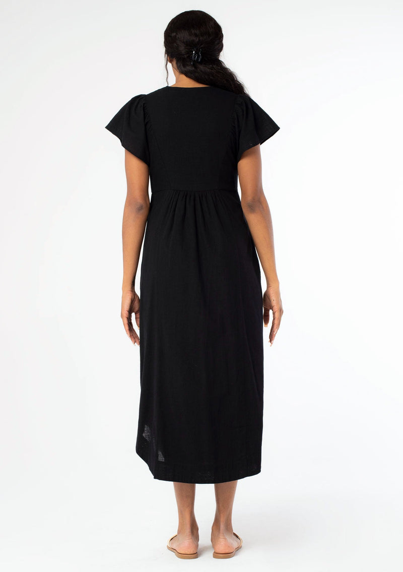 [Color: Black] A back facing image of a black woman wearing a black cotton mid length dress with a smocked top, button front skirt, and a loose, relaxed fit.