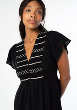 [Color: Black] A close up image of a black woman wearing a black cotton mid length dress with a smocked top, button front skirt, and a loose, relaxed fit.