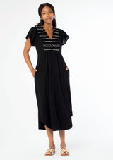 [Color: Black] A front facing image of a black woman wearing a black cotton mid length dress with a smocked top, button front skirt, and a loose, relaxed fit.