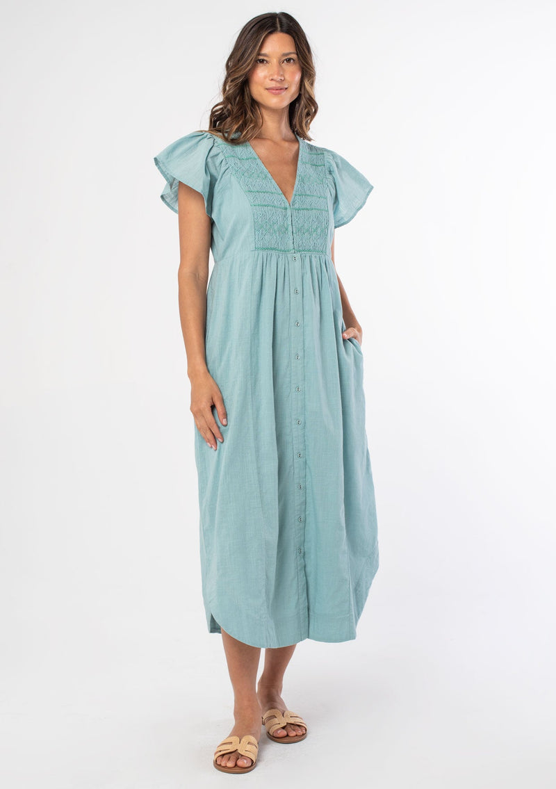 [Color: Light Aqua] A woman wearing a light aqua cotton mid length dress with a smocked top, button front skirt, and a loose, relaxed fit. 