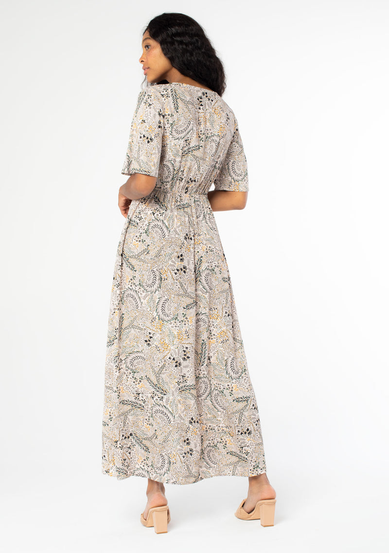 [Color: Natural/Olive] A back facing image of a black model wearing a natural and olive green floral print maxi dress, with short sleeves, a faux wrap front, and a front slit.