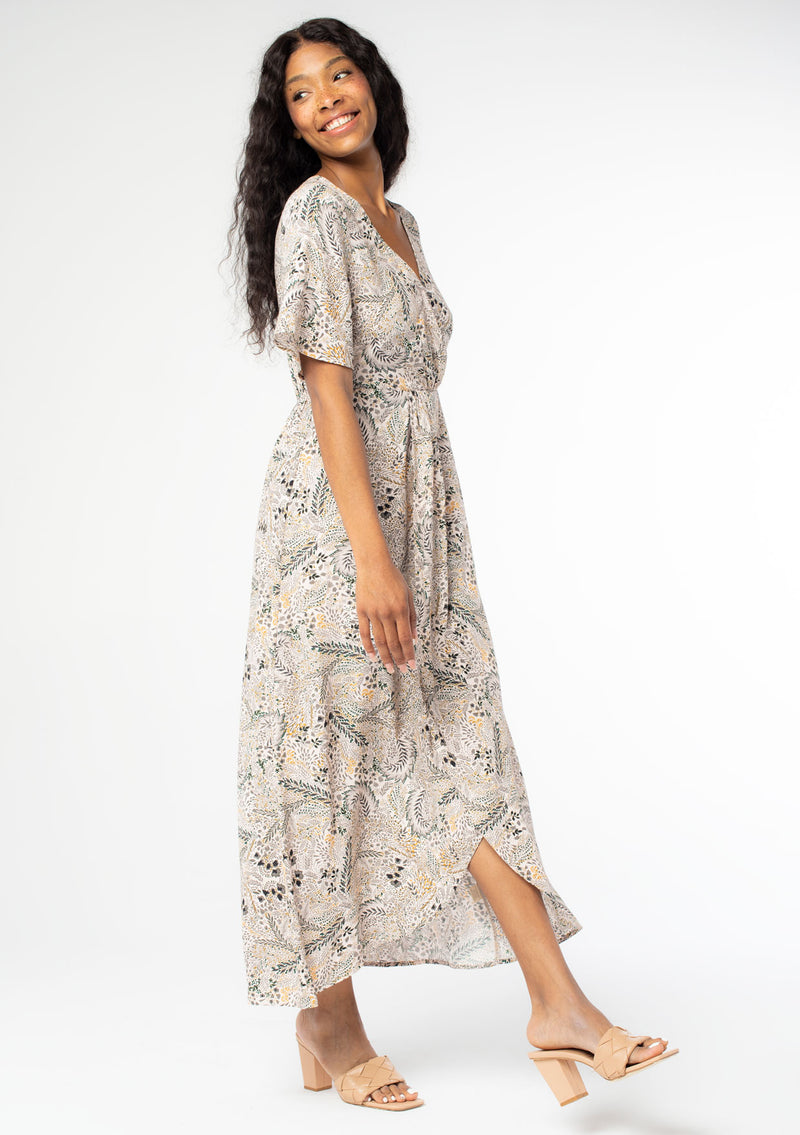 [Color: Natural/Olive] A side facing, full body image of a black model wearing a natural and olive green floral print maxi dress, with short sleeves, a faux wrap front, and a front slit.