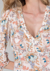 [Color: Natural/Coral] A close up front facing image of a blonde model wearing a bohemian spring mid length dress in a natural and coral floral print. With half length sleeves, tie cuffs, side slits, and a v neckline. 