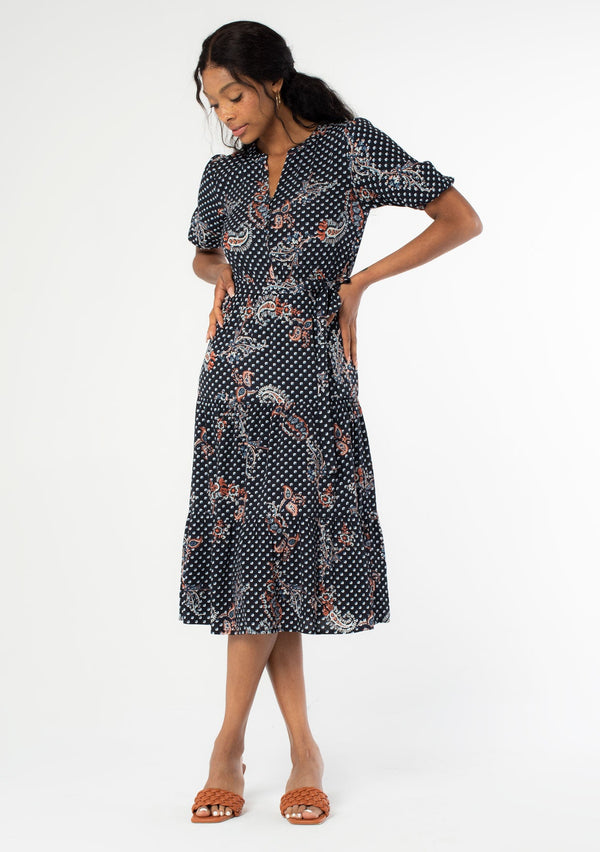 [Color: Charcoal/Dusty Blue] A full body front facing image of a black model wearing a charcoal dark grey mid length dress with blue paisley dot print throughout. A bohemian midi dress with short puff sleeves, a flowy tiered skirt, and a self tie waist belt. 