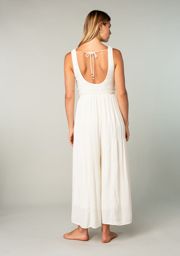 [Color: Ivory] A back facing image of a blonde model wearing an ivory white sleeveless one piece jumpsuit with a crochet knit top, a wide pant leg, a scooped neckline, side pockets, and an open back with tie closure.