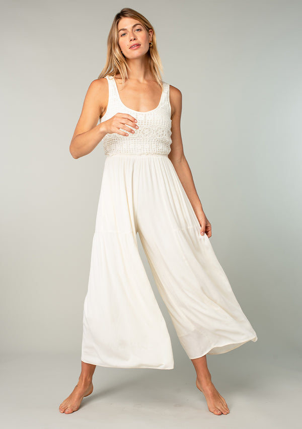 [Color: Ivory] A front facing image of a blonde model wearing an ivory white sleeveless one piece jumpsuit with a crochet knit top, a wide pant leg, a scooped neckline, side pockets, and an open back with tie closure.